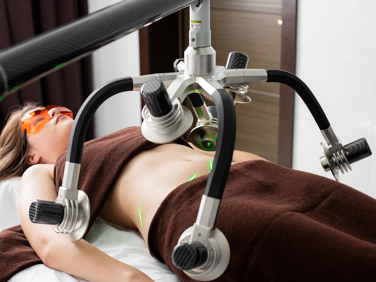 Female client lying on table under lasers of modern weight loss machine on belly during liposuction procedure in medical clinic